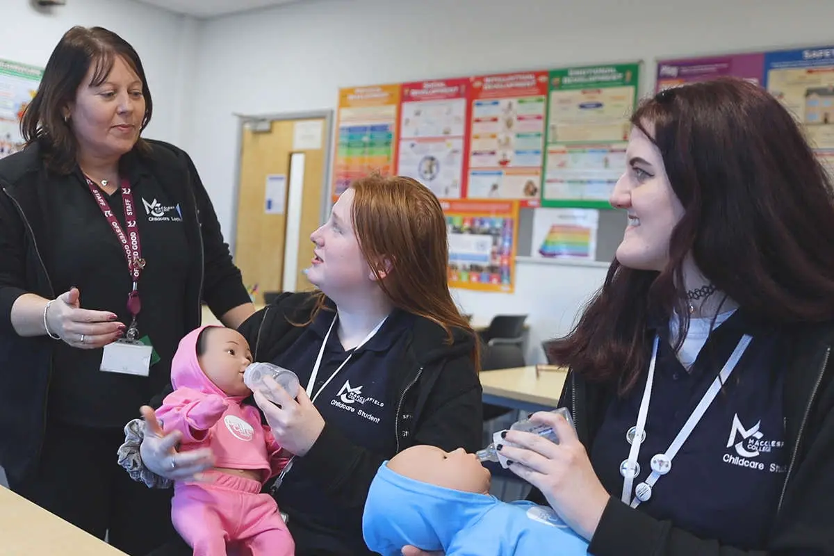 2 childcare students happily practice giving bottles to dolls while the tutor observes