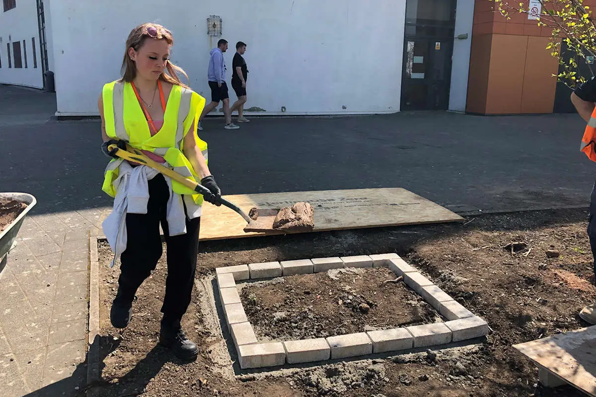 a female students with a spade and cement walks past a partly built wall