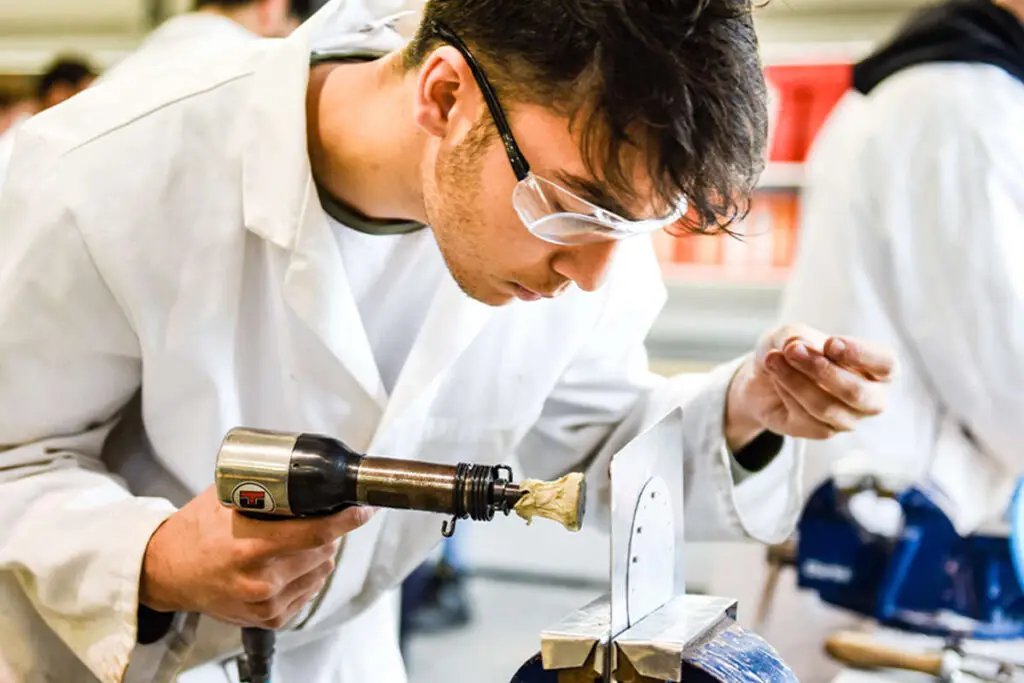 A student in a white lab coat and safety glasses using a heat gun