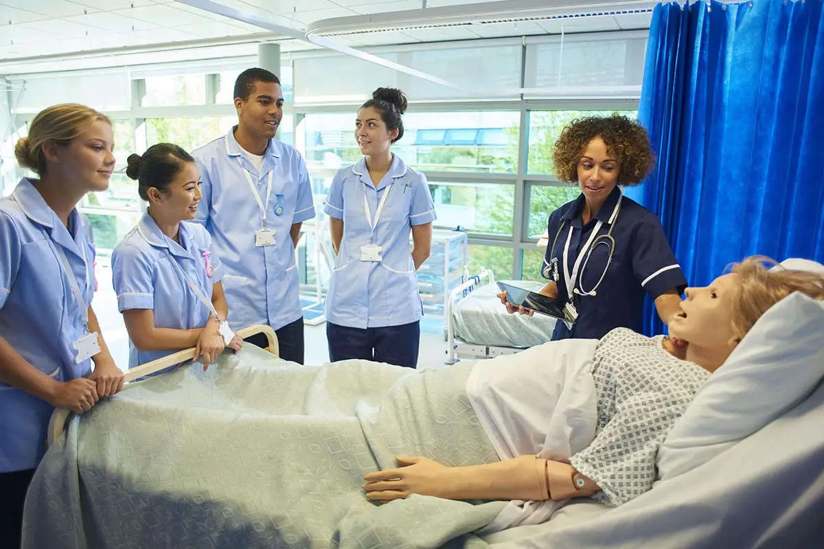 a group of student nurses stand around a hospital bed with a dummy patient in