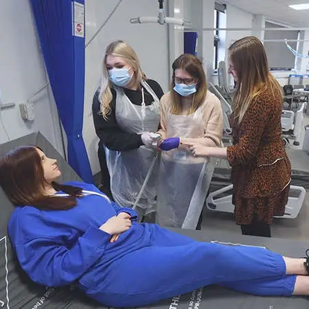 2 health care students adjust a hospital bed while another student lies on top of it. A tutor observes