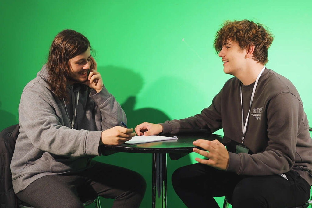 2 media students chatting happily while sat at a table in front of a green screen