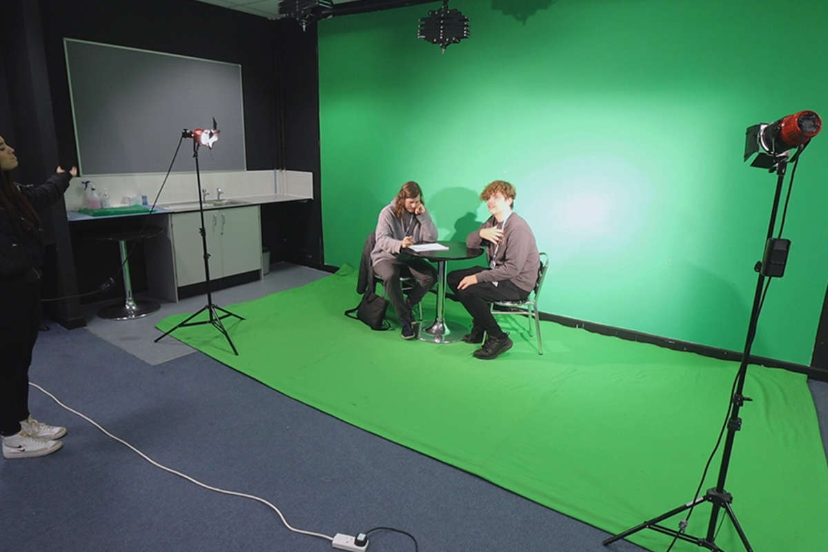 2 students sat at a table in front of a green screen