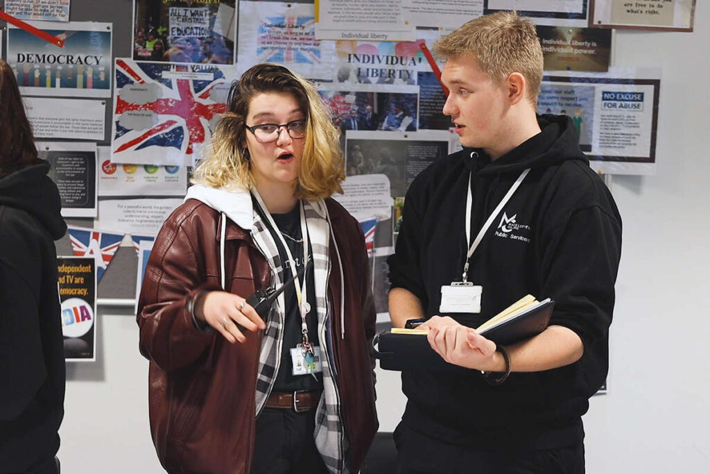 2 people talking, one is holding a walkie talkie, the other is holding a note book