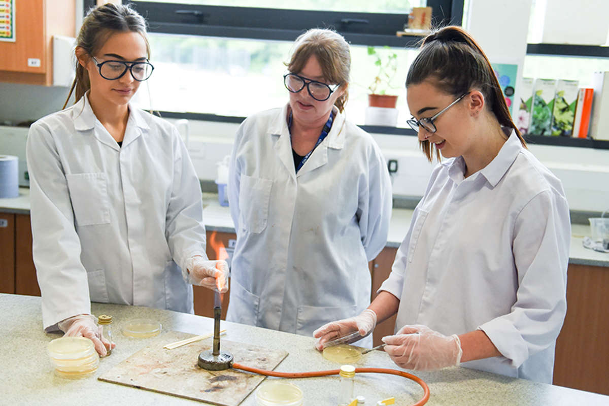 2 students and a tutor standing behind a lit bunsen burner, inspecting the contents of petri dishes