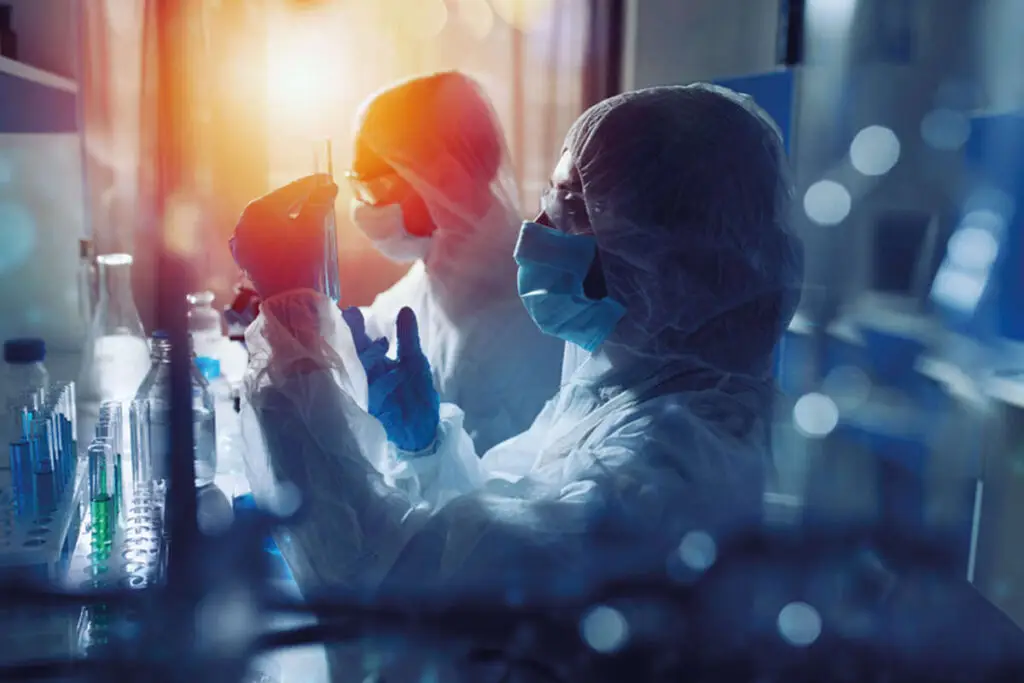 2 people in science lab, wearing white overalls, gloves, masks and safety glasses inspect blue liquid in a test tube