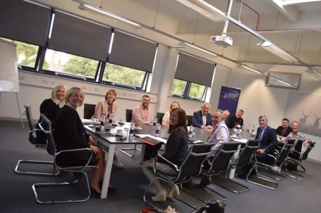 Staff members are sat round a table during a business meeting. They are smiling for a photo.