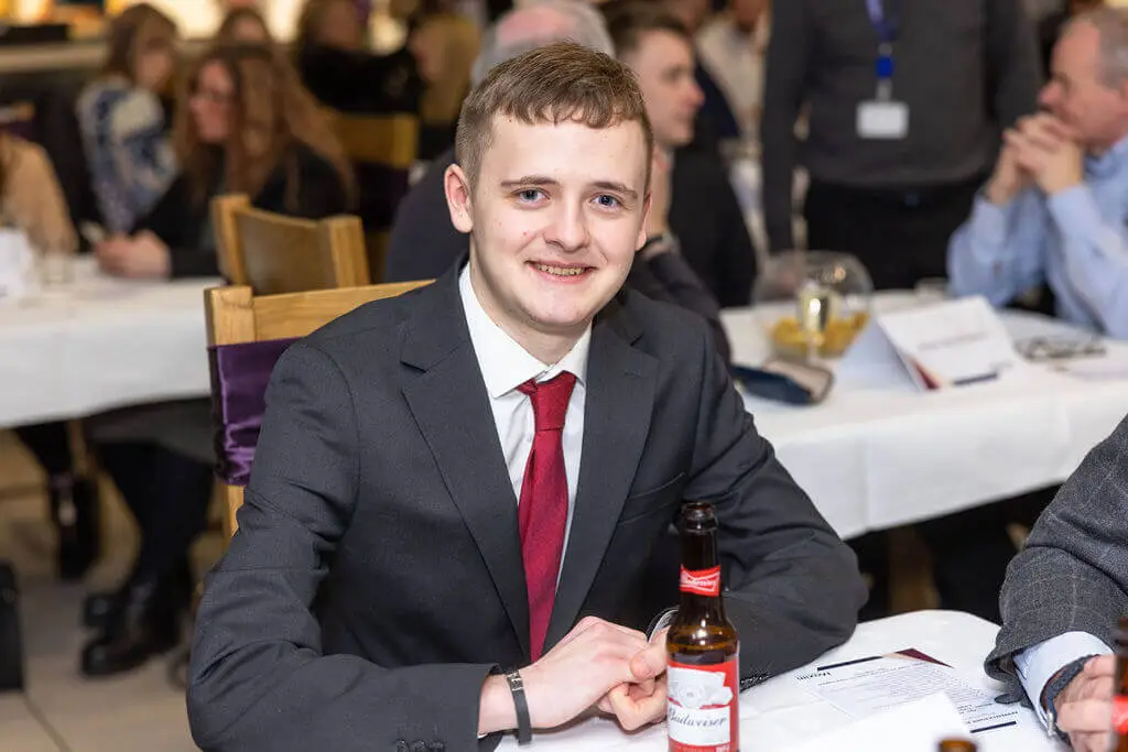 A male student is sitting at a table during an award's ceremony. He is smiling for a photo.