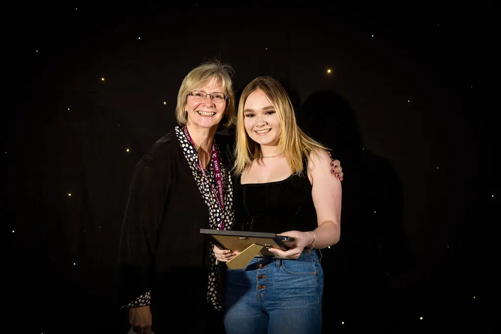 A student and a staff member are smiling for a photo during an award ceremony. The student is holding an award