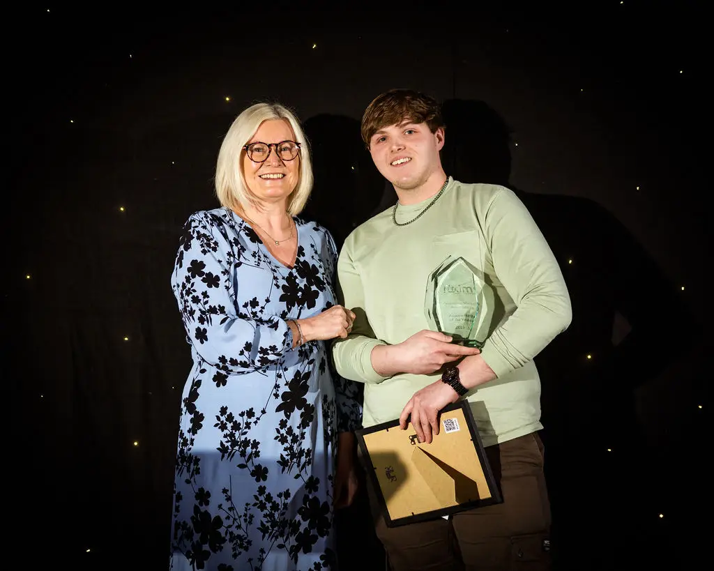 A student and a staff member are smiling for a photo during an award ceremony. The student is holding a glass award and a certificate