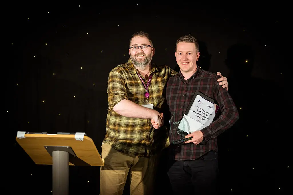 A student and a staff member are smiling for a photo during an award ceremony. The student is holding a certificate and a glass award