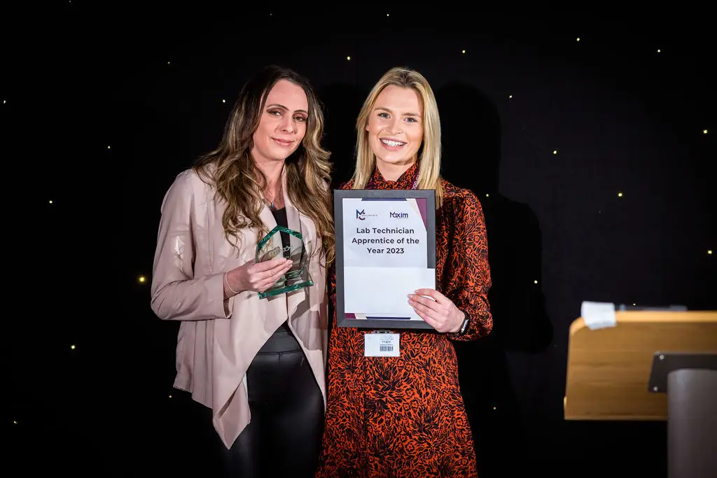 A student and a staff member are smiling for a photo during an award ceremony. The student is holding a glass award while the staff member is holding a certificate