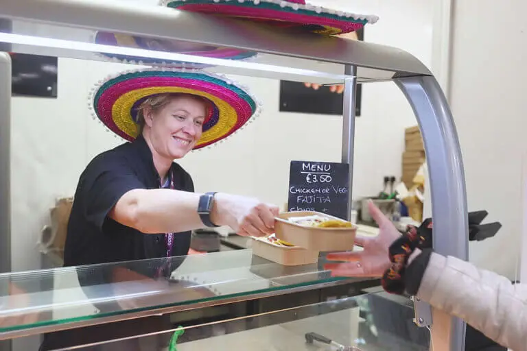 A staff member is wearing a sombrero hat and is serving food in the canteen