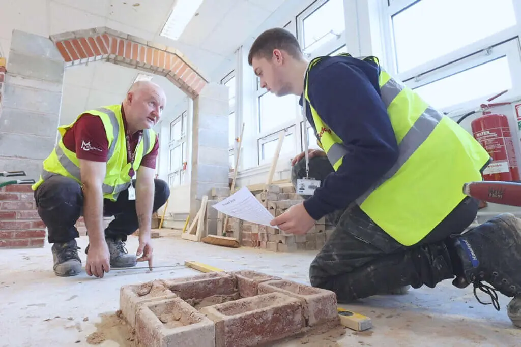 A construction student and teacher are setting out bricks. The student is holding a white piece of paper in his hand.