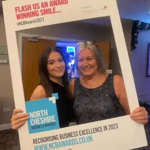 Two staff members are smiling for a photo whilst holding a white cardboard sign that says north Cheshire business awards