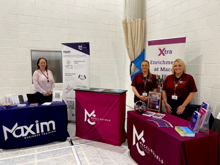 Three staff members are smiling for a photo during a school's event at Macclesfield College. In front of them, there are tables filled with flyers and sweets.