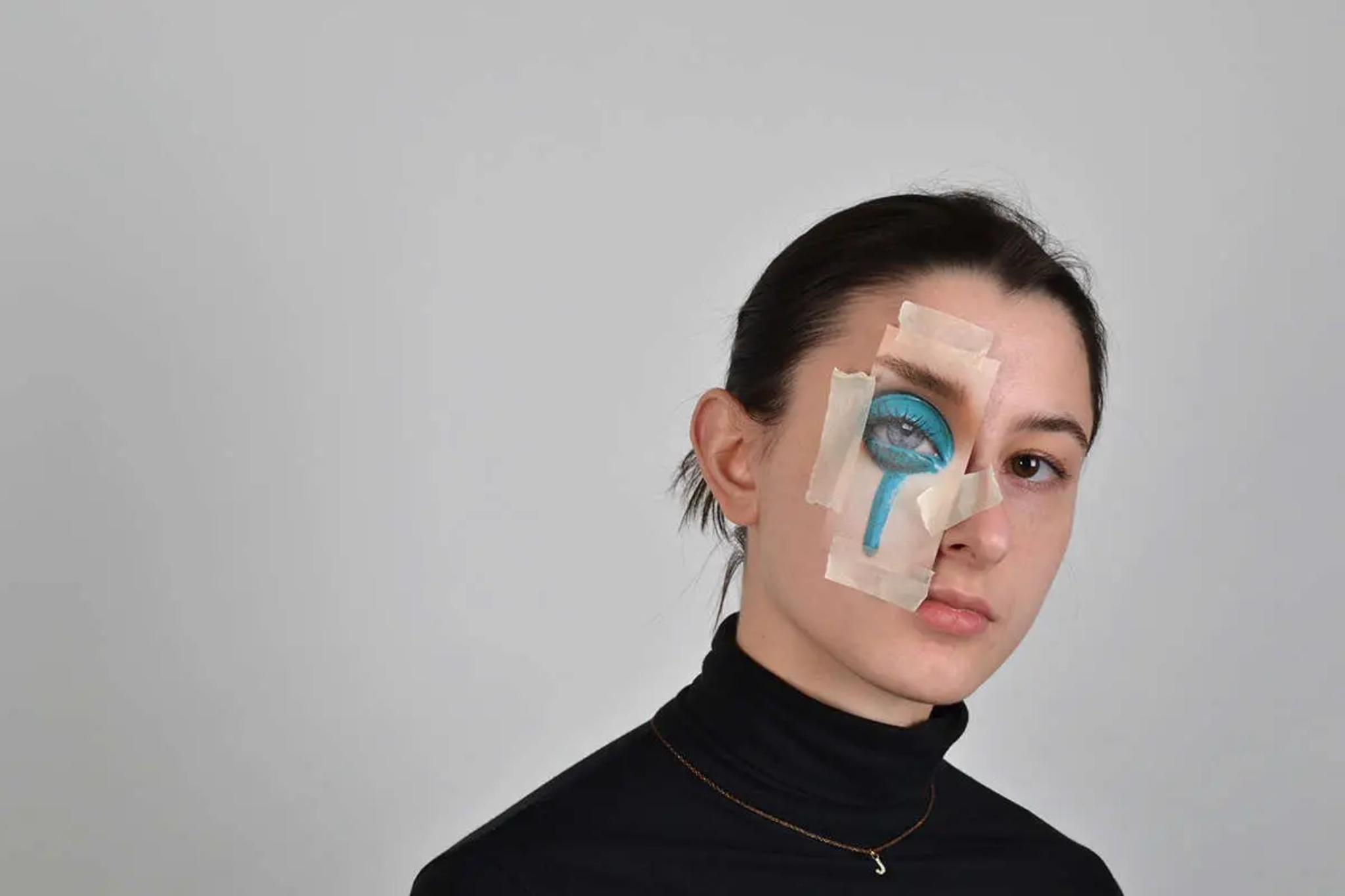 A woman in a black turtle neck has a drawing of an eye taped over one of her eyes. The drawing has bright blue eyeshadow over the eye lid and is running down the cheek