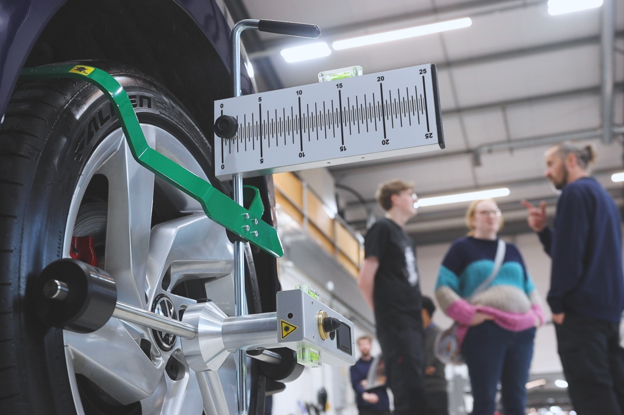 Measuring equipment is attached to a car wheel and tyre. Near the equipment there are two visitors and an Automotive teacher talking together.