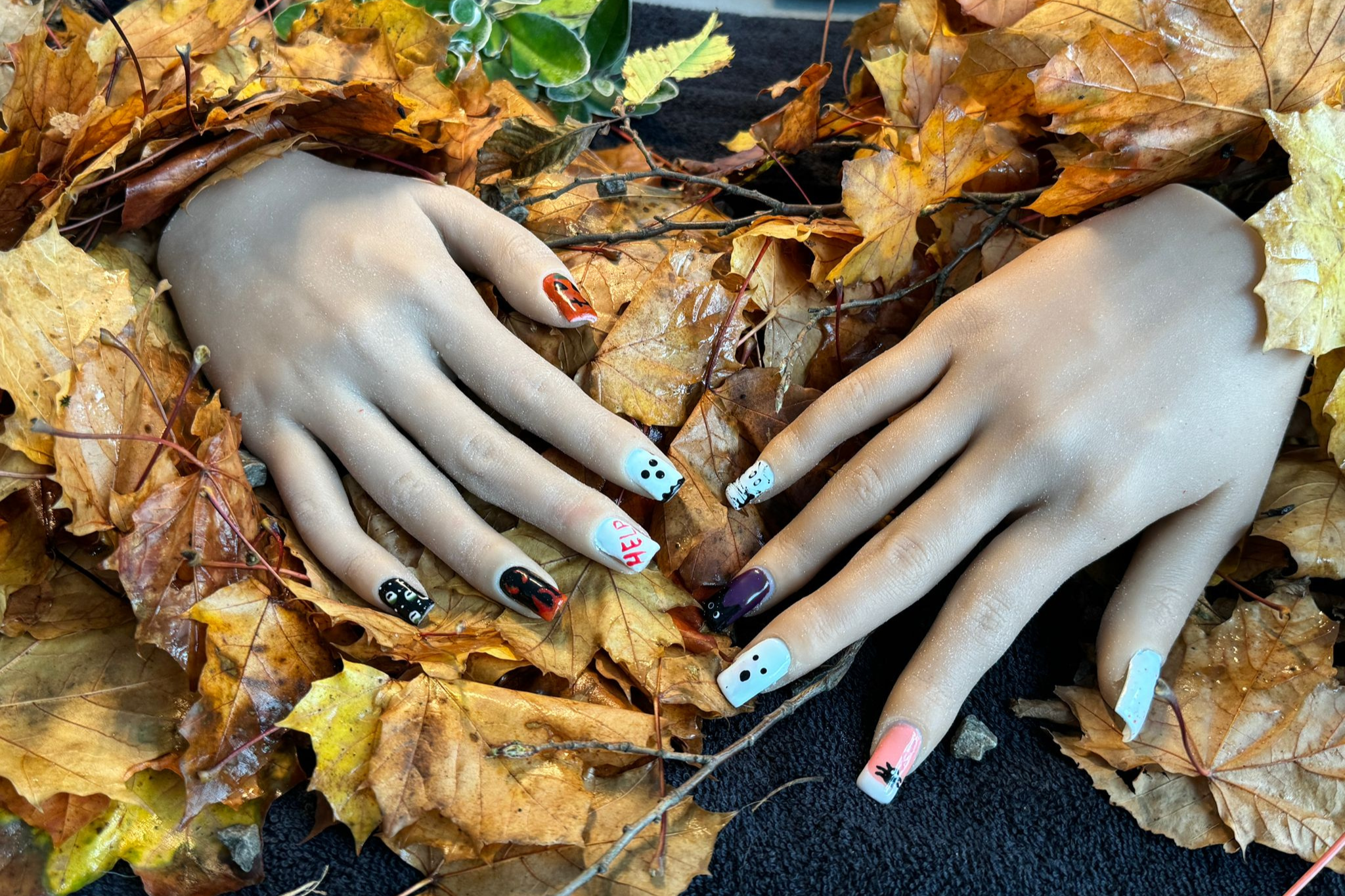 Two mannequin hands that are with brightly coloured nails painted by beauty students lie under autumn leaves.