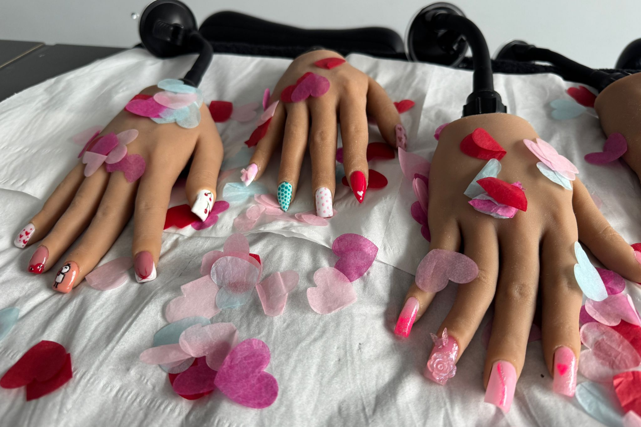 A table is filled with three mannequin hands containing brightly coloured nails by Beuaty Students. Next to the hands, there are paper hearts decorating the table.