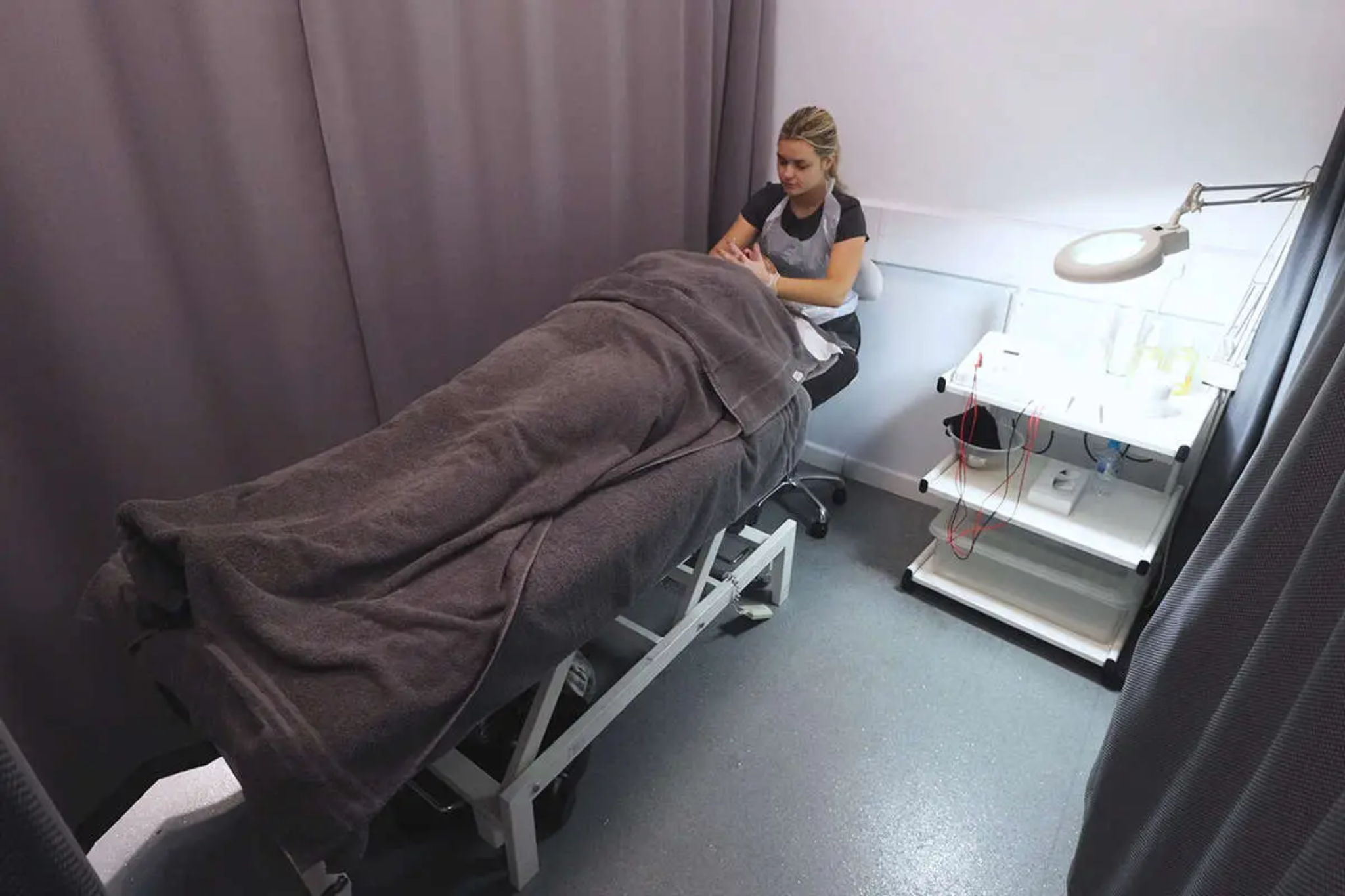 A beauty student sits down while giving someone a facial treatment. The person receiving the treatment is on a bed and covered with towels