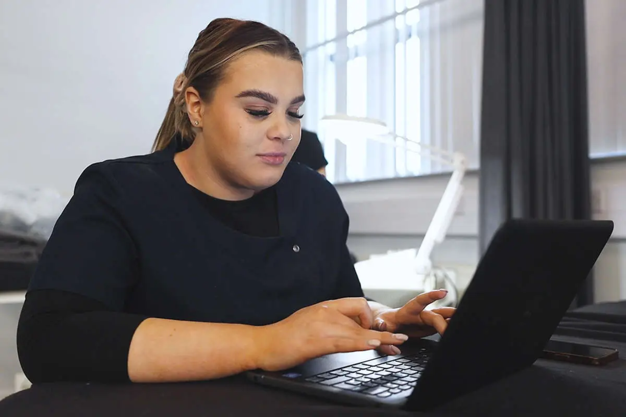 A beauty student, dressed in a smart navy blue uniform, is working on a laptop