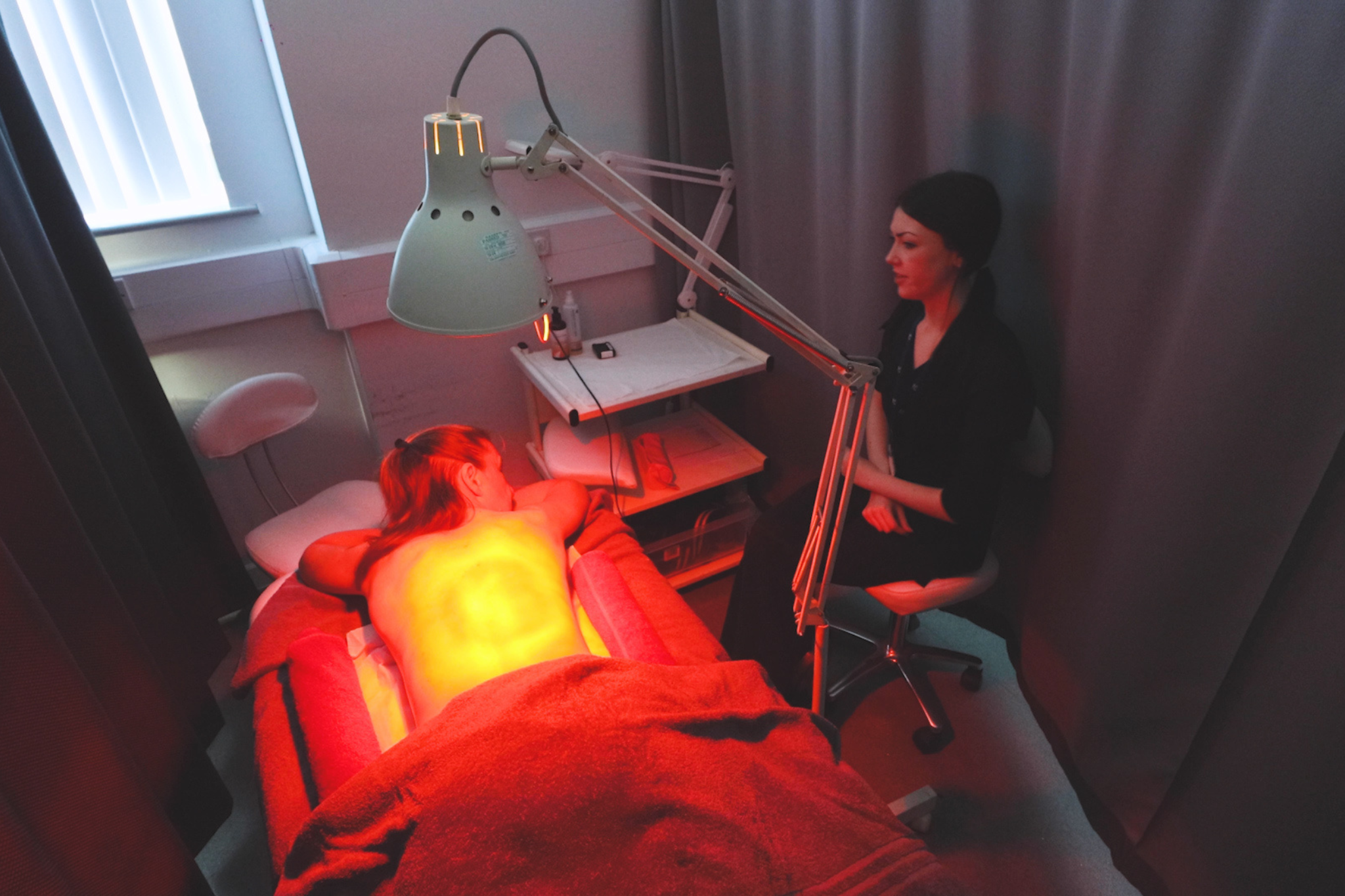 A client lies comfortably on a treatment bed with a red light shining on their back, as a beauty student prepares to begin the treatment.