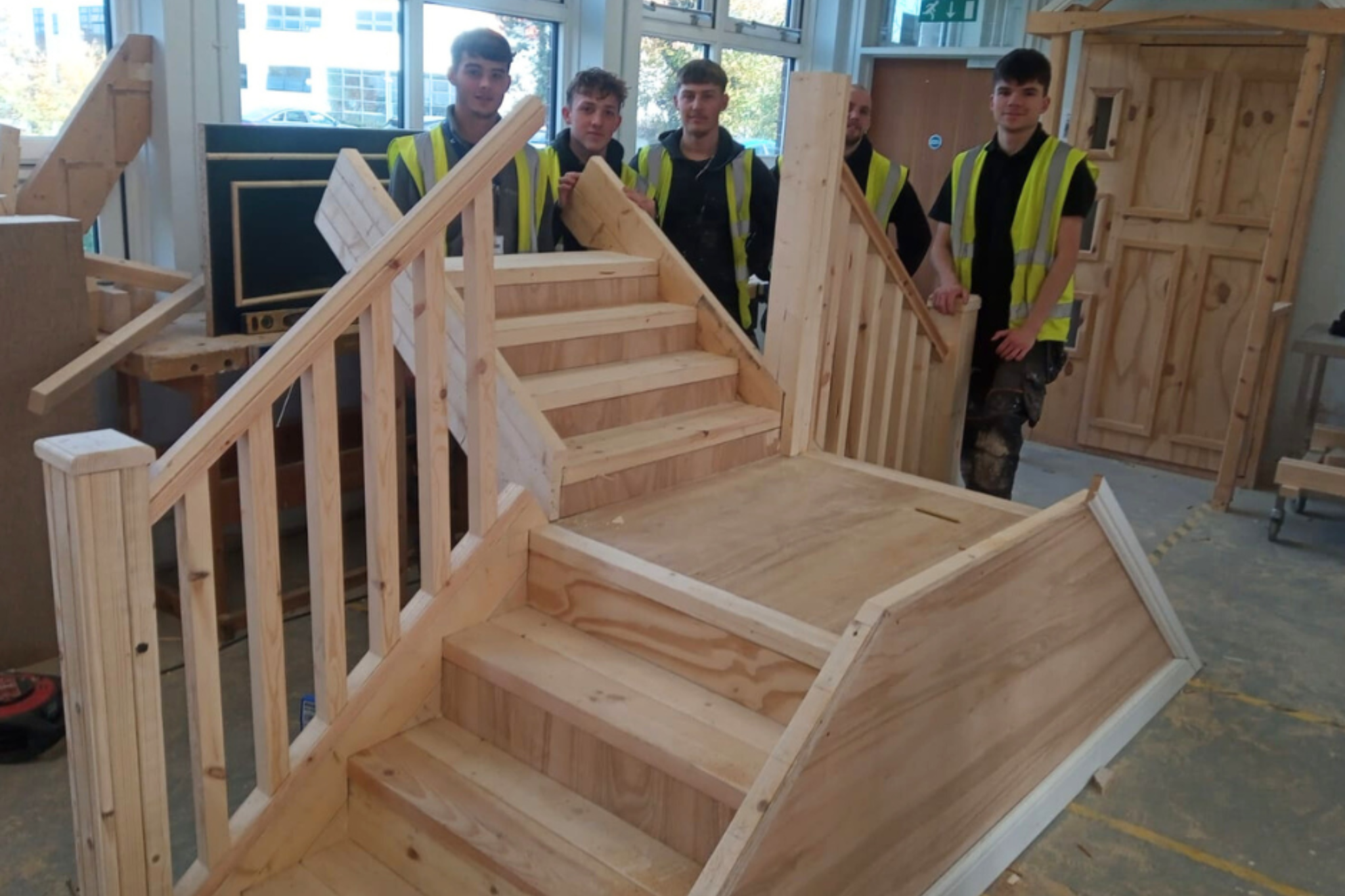 A group of Carpentry & Joinery apprentices smiling for a photo, standing next to the wooden staircase they have built.