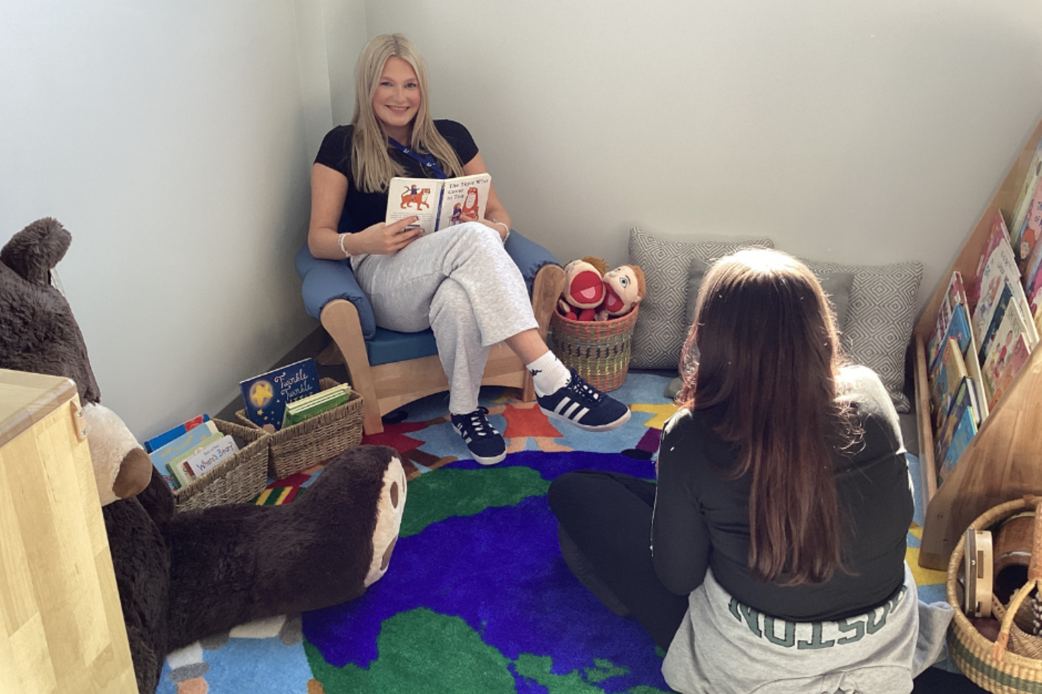 Childcare students are inside Macclesfield College's Skills Hub Nursery. One student is sitting in a chair reading a book.