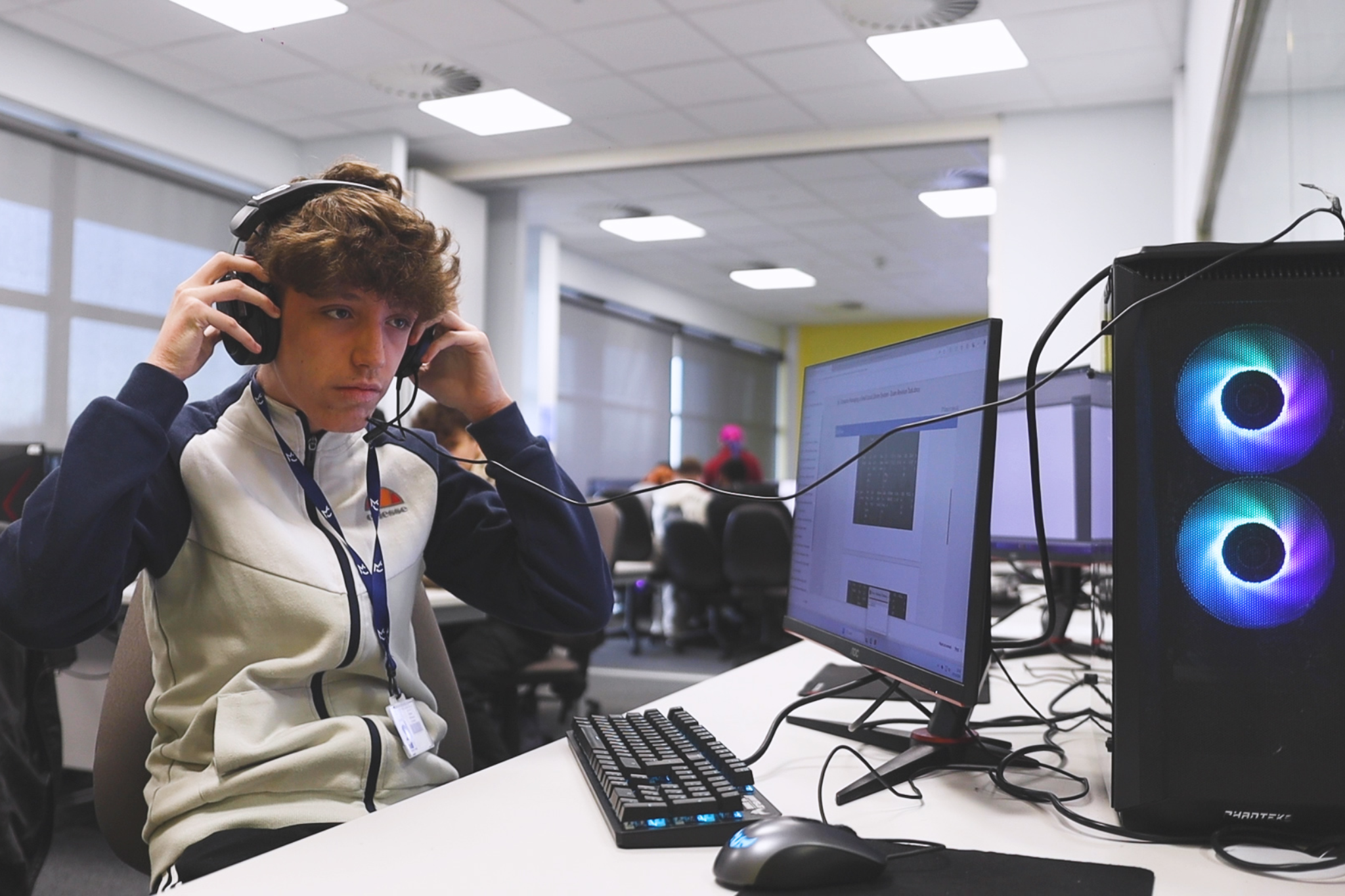 A Computing student is putting on headphones whilst they work at a computer.