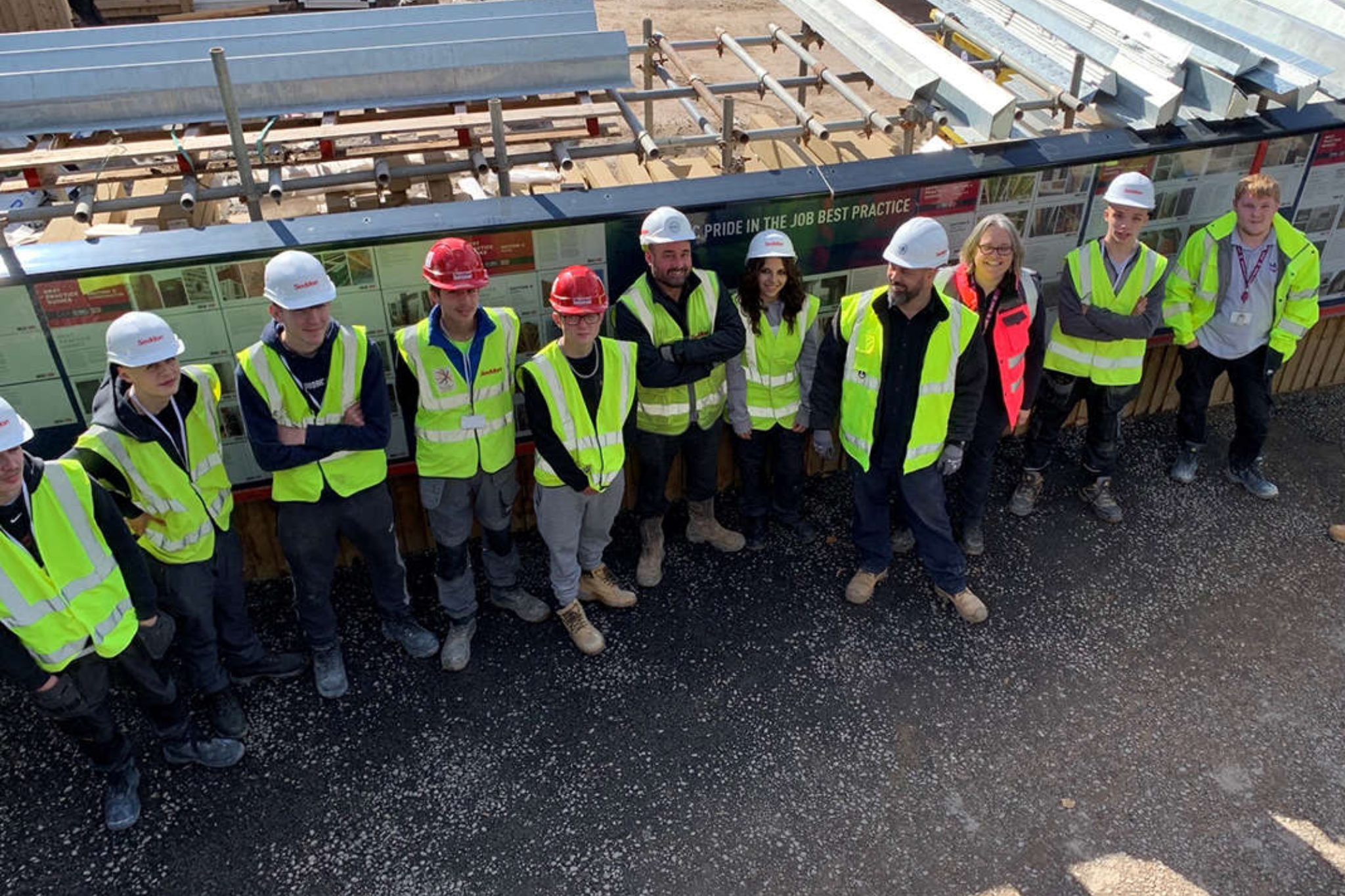 A group of students and staff members are standing together on a construction site, smiling for a photo. The students and staff members are all wearing PPE and construction uniform.