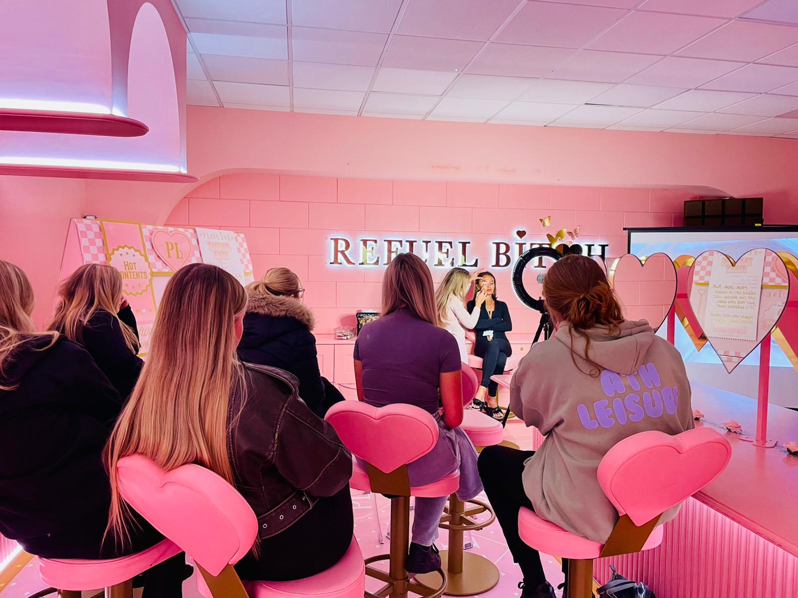 A group of students are sitting on pink heart chairs while they watch a beauty demonstration from PLouise Cosmetics