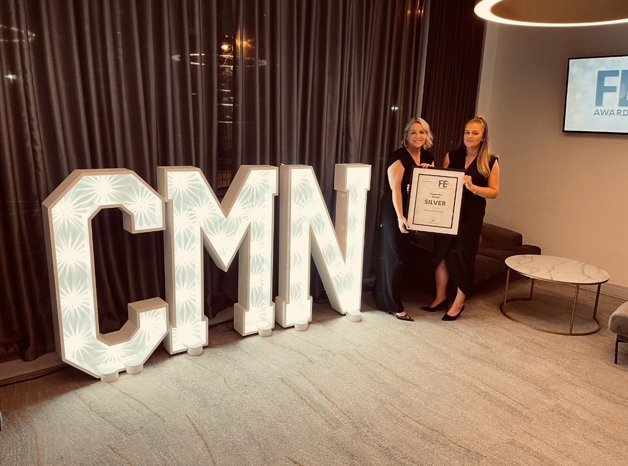 Two female staff members from Macclesfield College are holding a framed certificate and smiling for a photo. Next to them are large illuminated letters that read CMN