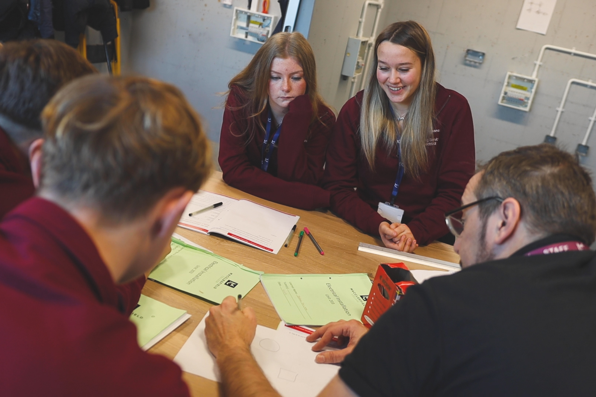 A group of electrical students are standing around a workshop bench, watching their teacher talk about a drawing.