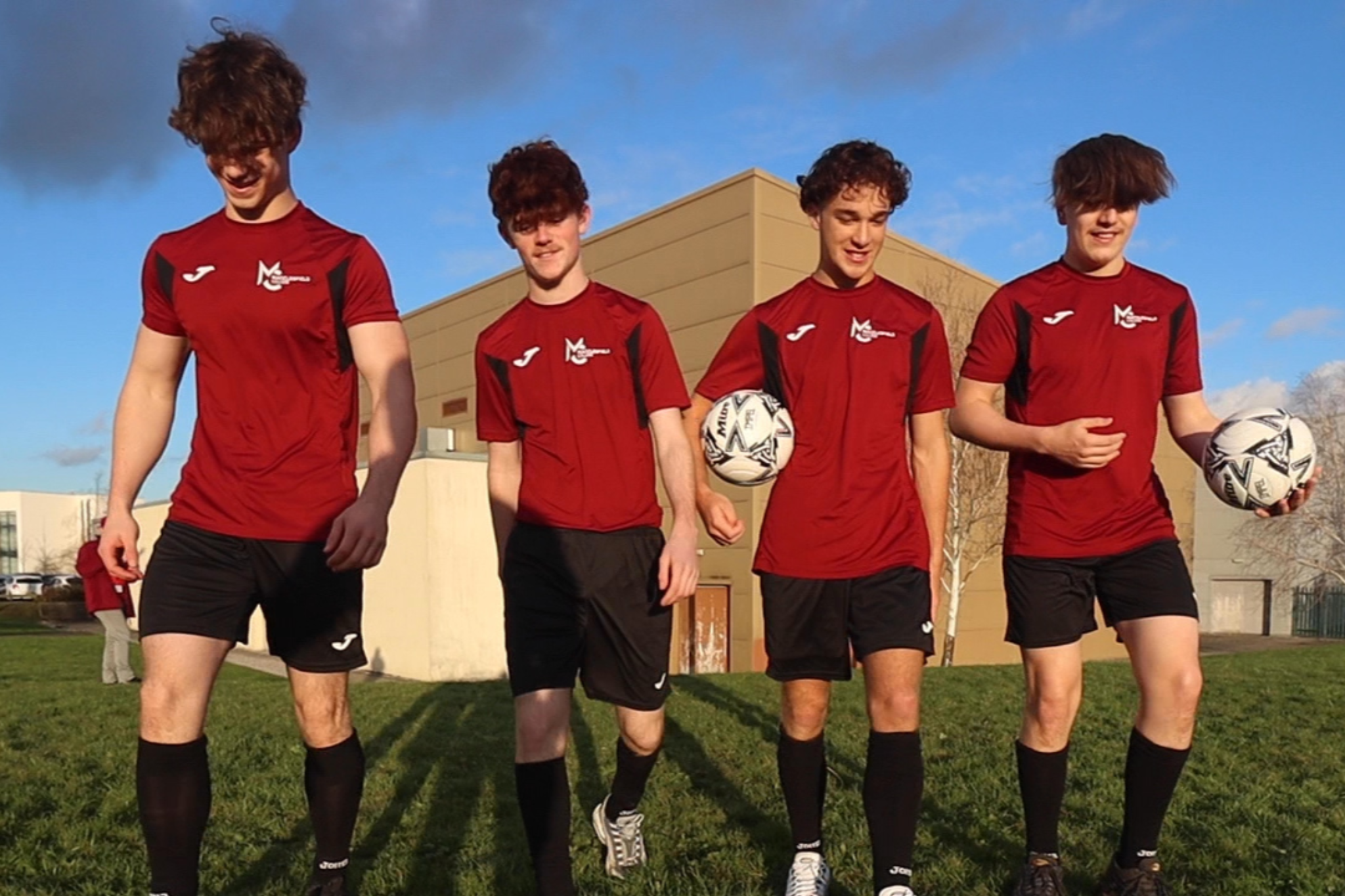 Four sports students are walking on green grass. One student is holding a football