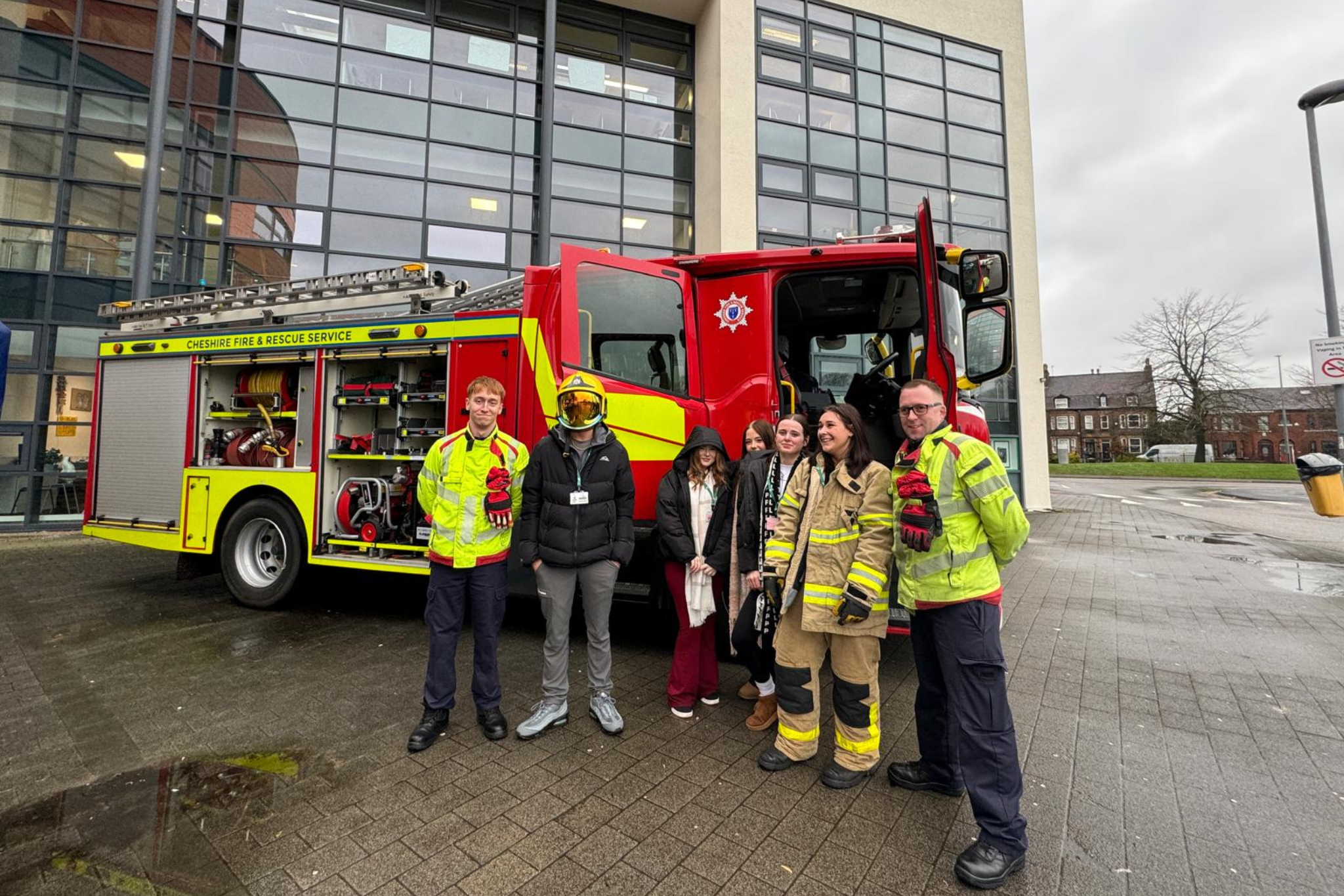 A group of students and firefighters stand smiling for a photo by a fire truck