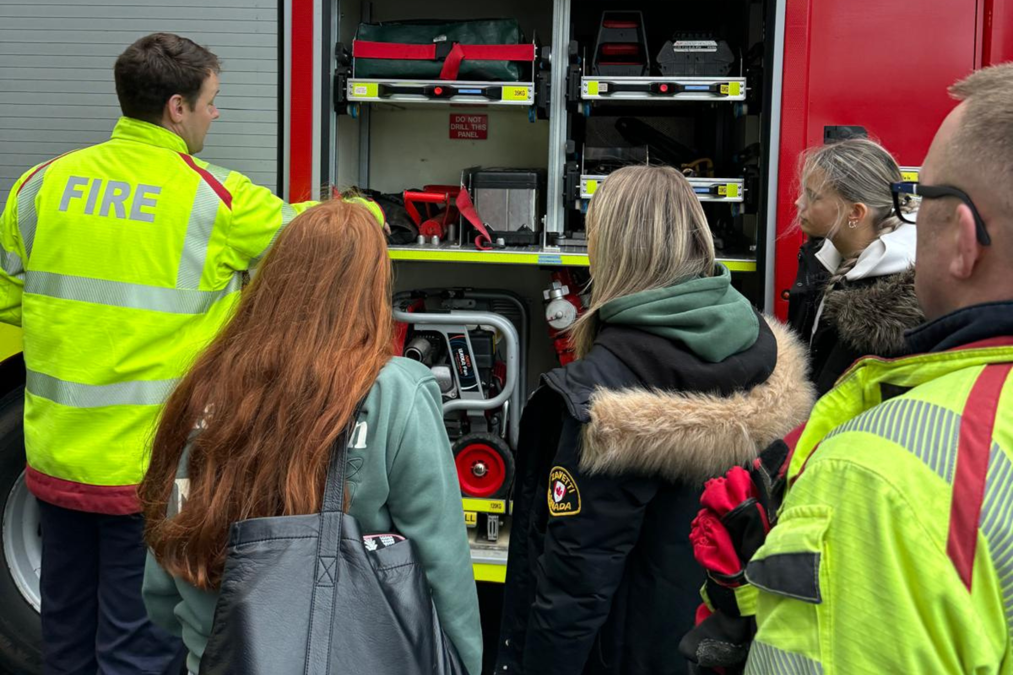 A group of students and two firefighters are looking at a fire truck