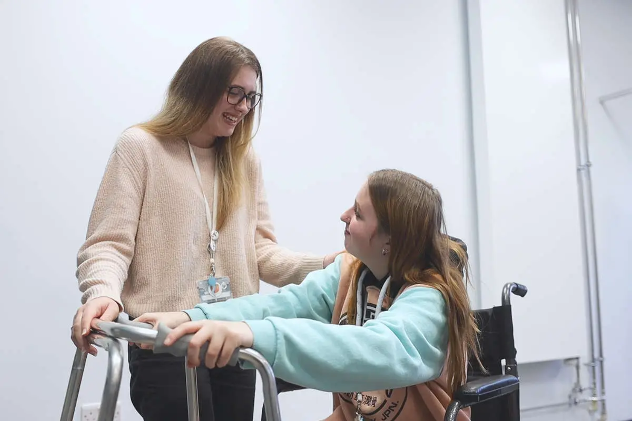 The image shows two health students in Macclesfield College's Health Hub. One student is sitting in a wheelchair using a hospital zimmer frame. While the other is standing next to them.