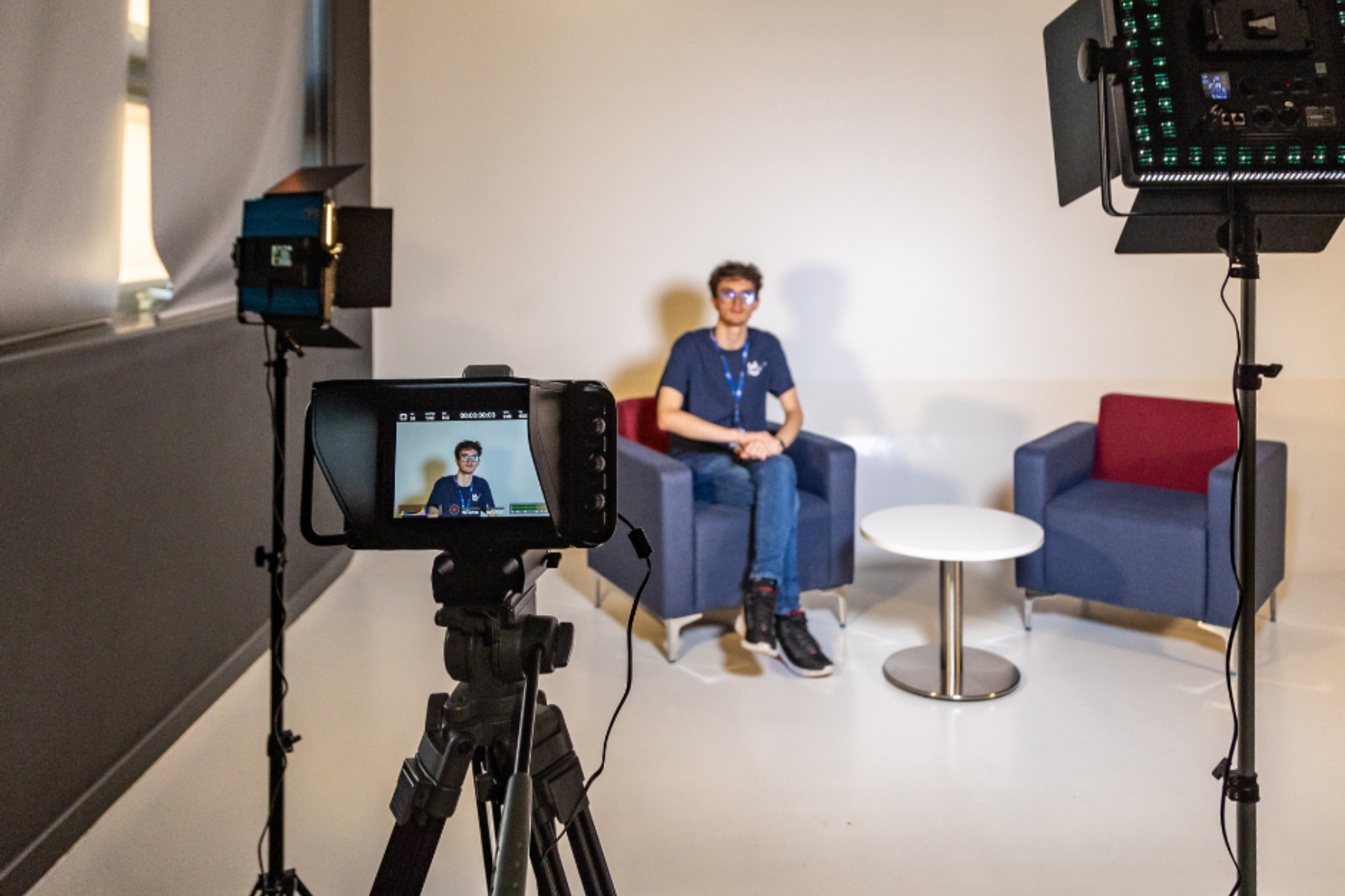 The photo shows an interview set-up with two chairs and a table, ready for filming. Lighting equipment and a camera are positioned to capture the scene and a Creative Media student is seated in one of the chairs.
