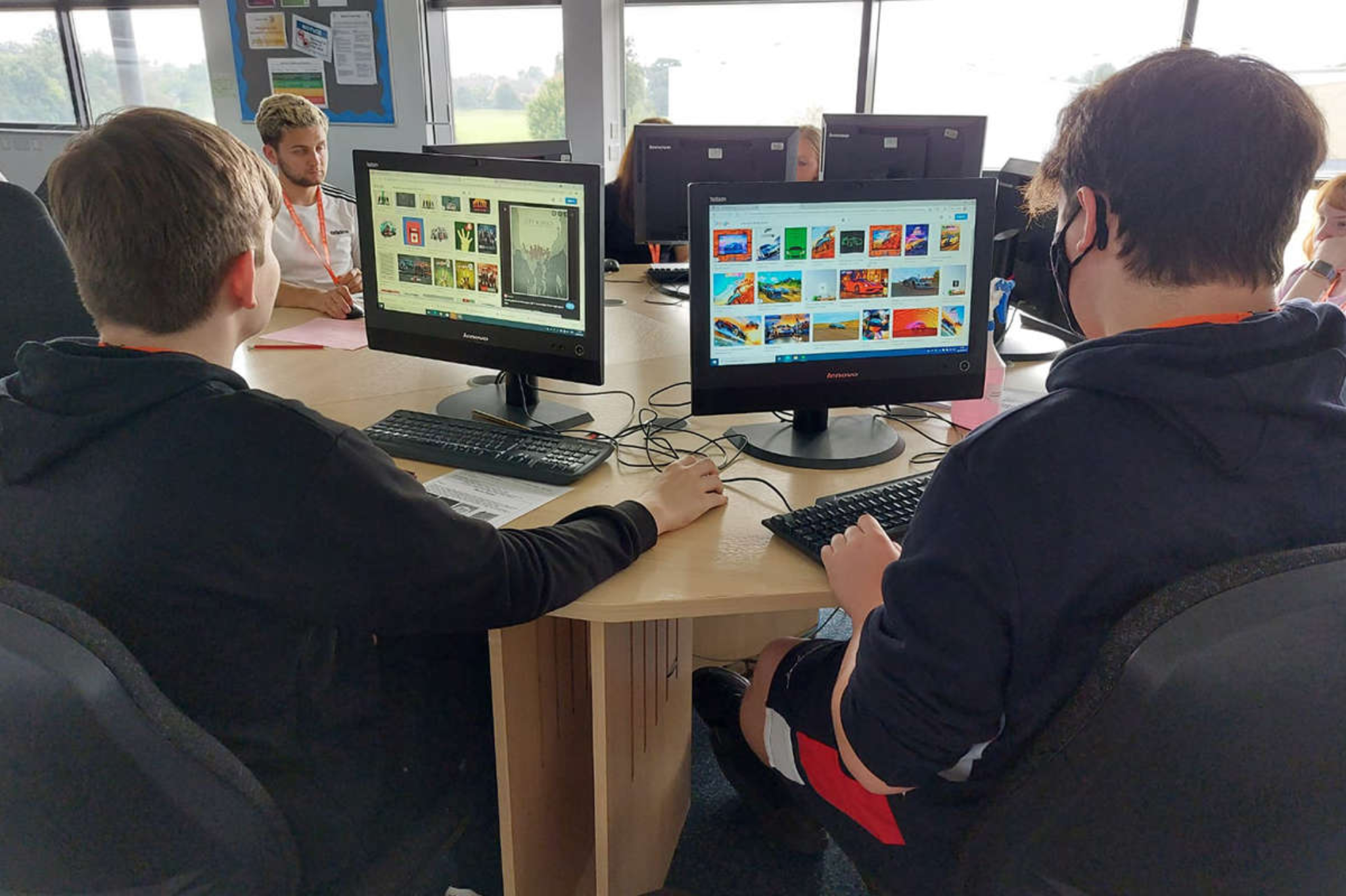 A group of students are sitting in front of computers at a large, round circular desk. On their computer screens, there are media files.
