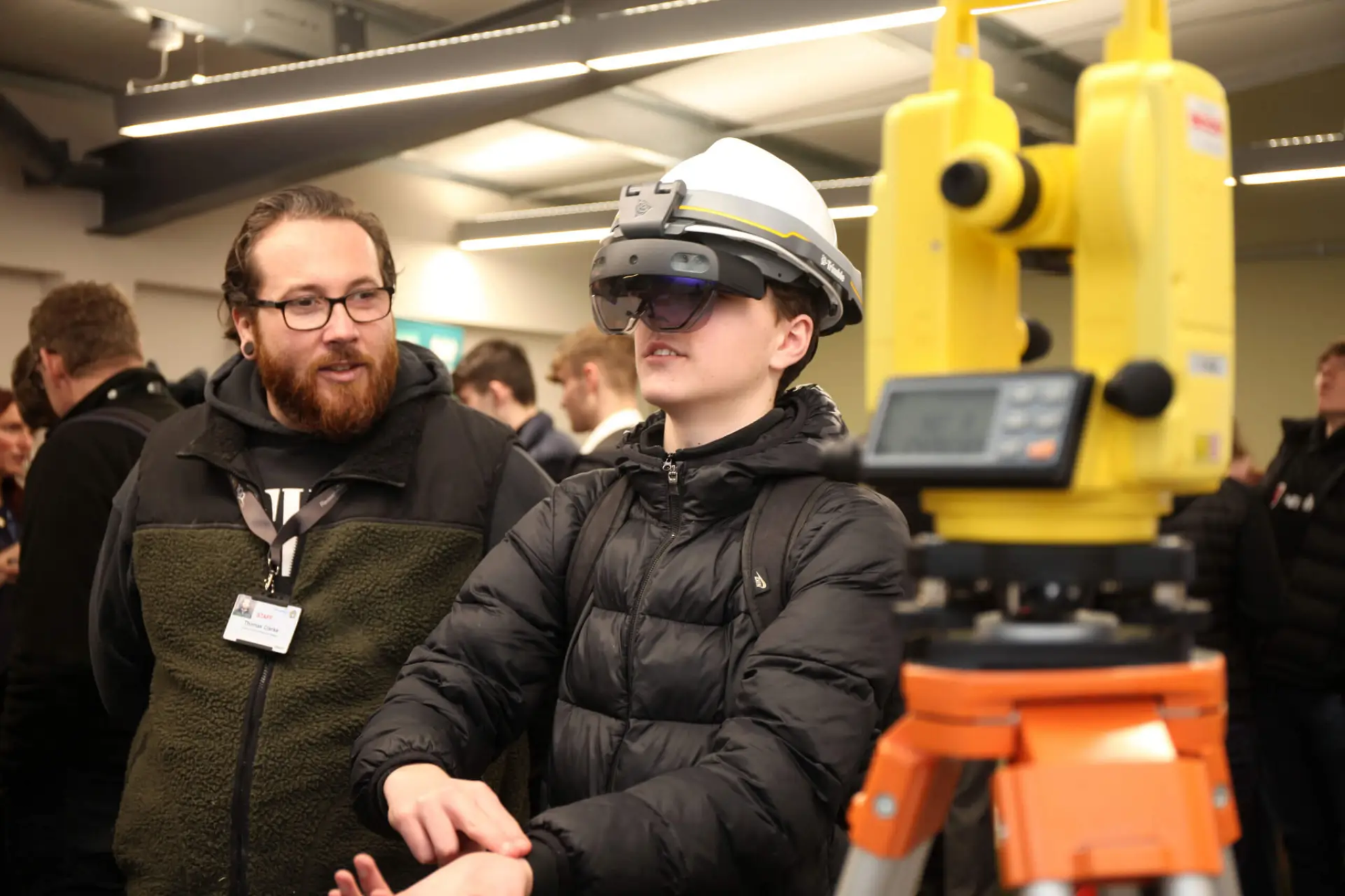 A student and a staff member stand next to each other. A male student is wearing a construction helmet and glasses. Next to them is surveying equipment