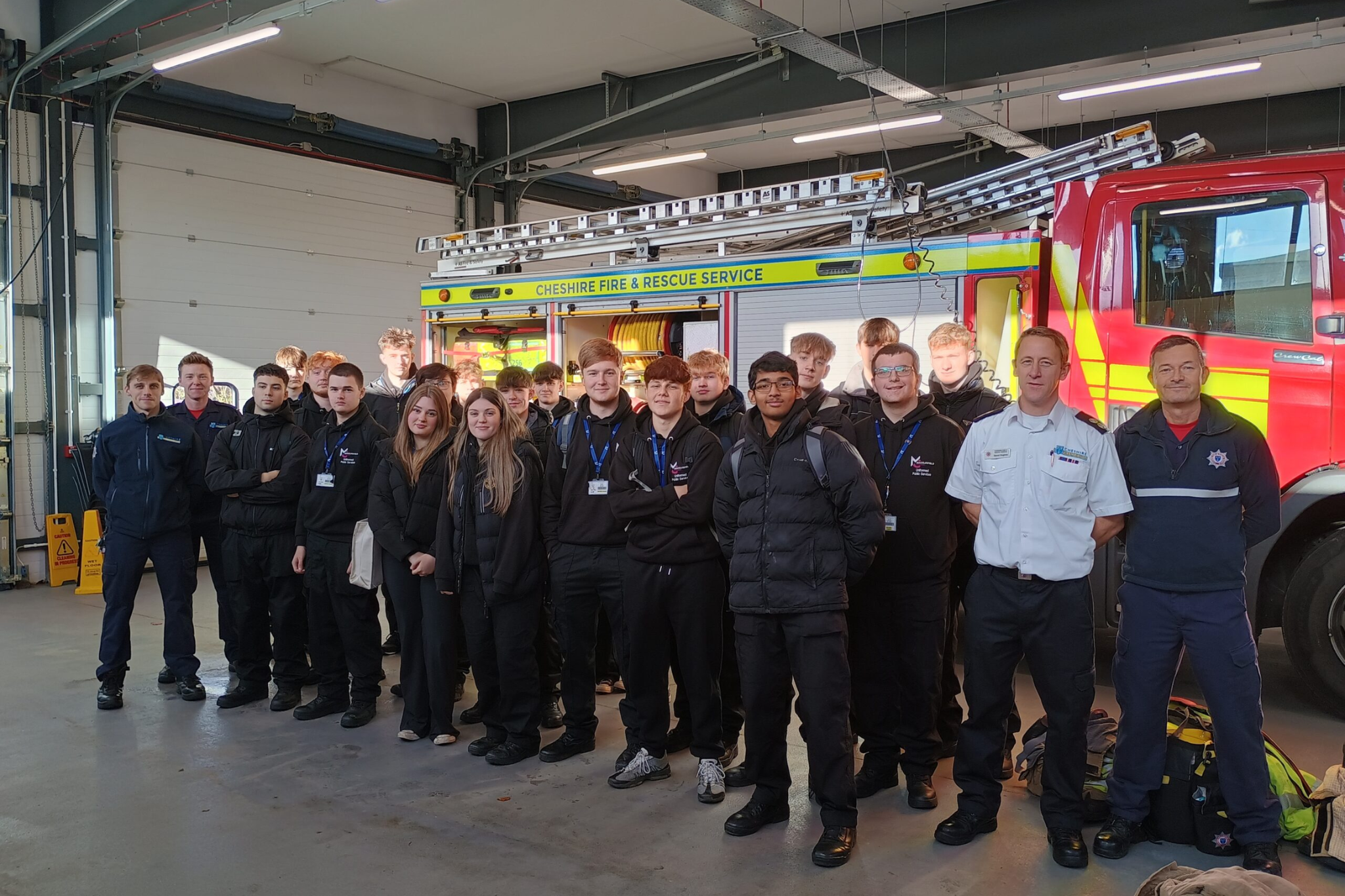 Public Service students are smiling with firemen as they pose for a photo. They are standing in front of red fire engine.