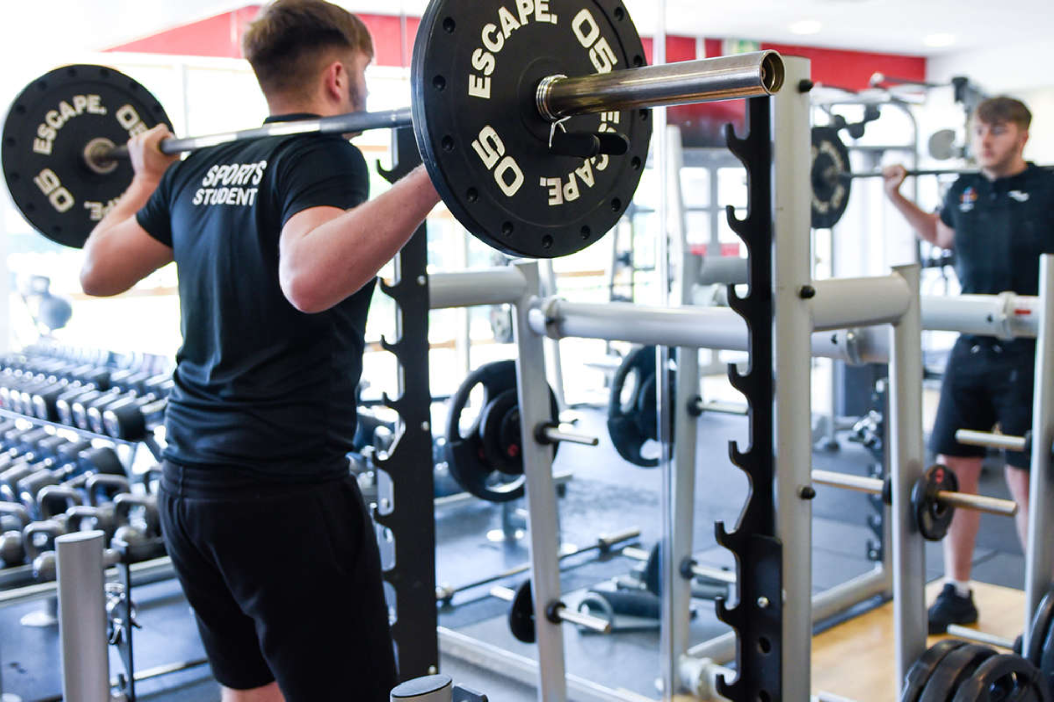 A young male holding a weight bar across the back of his shoulders while looking in the mirror