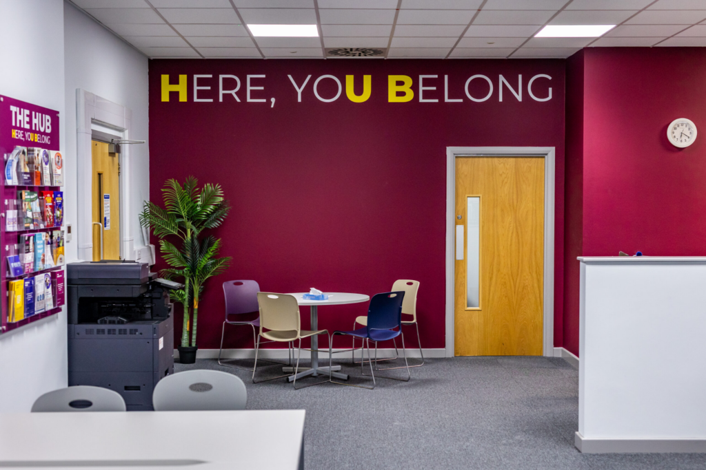A photo of Macclesfield College's Student Hub showing a printer, a noticeboard, and a table with chairs. A quote on the wall reads, “Here, You Belong.”