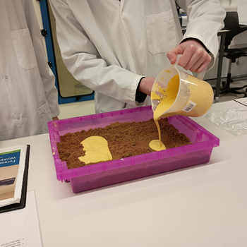 a student is pouring a jug of custard into a box filled with sand