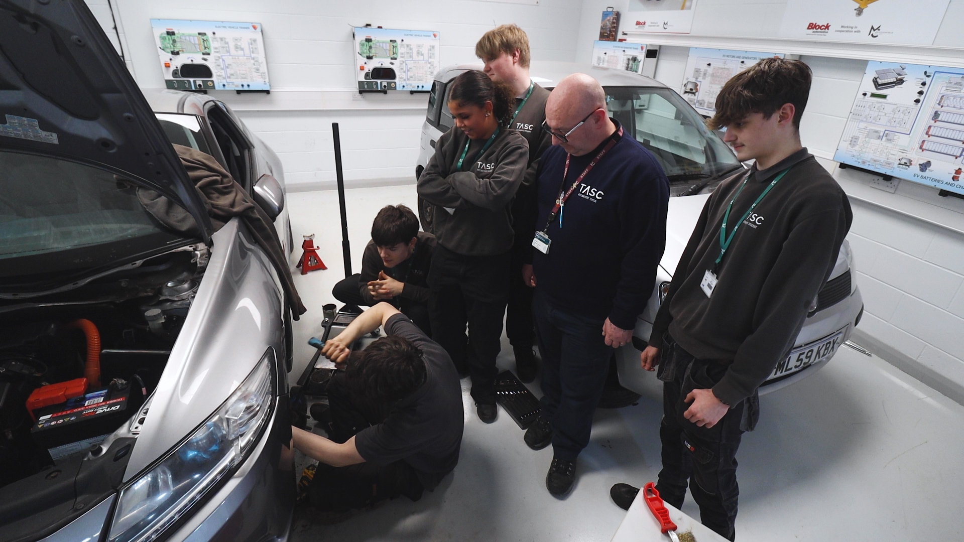 a group of students and a teacher watch as an automotive student changes a tyre