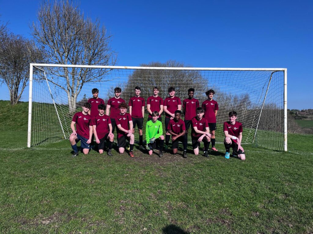 a group of students wearing football kits are sitting in front of goal, smiling for a photo