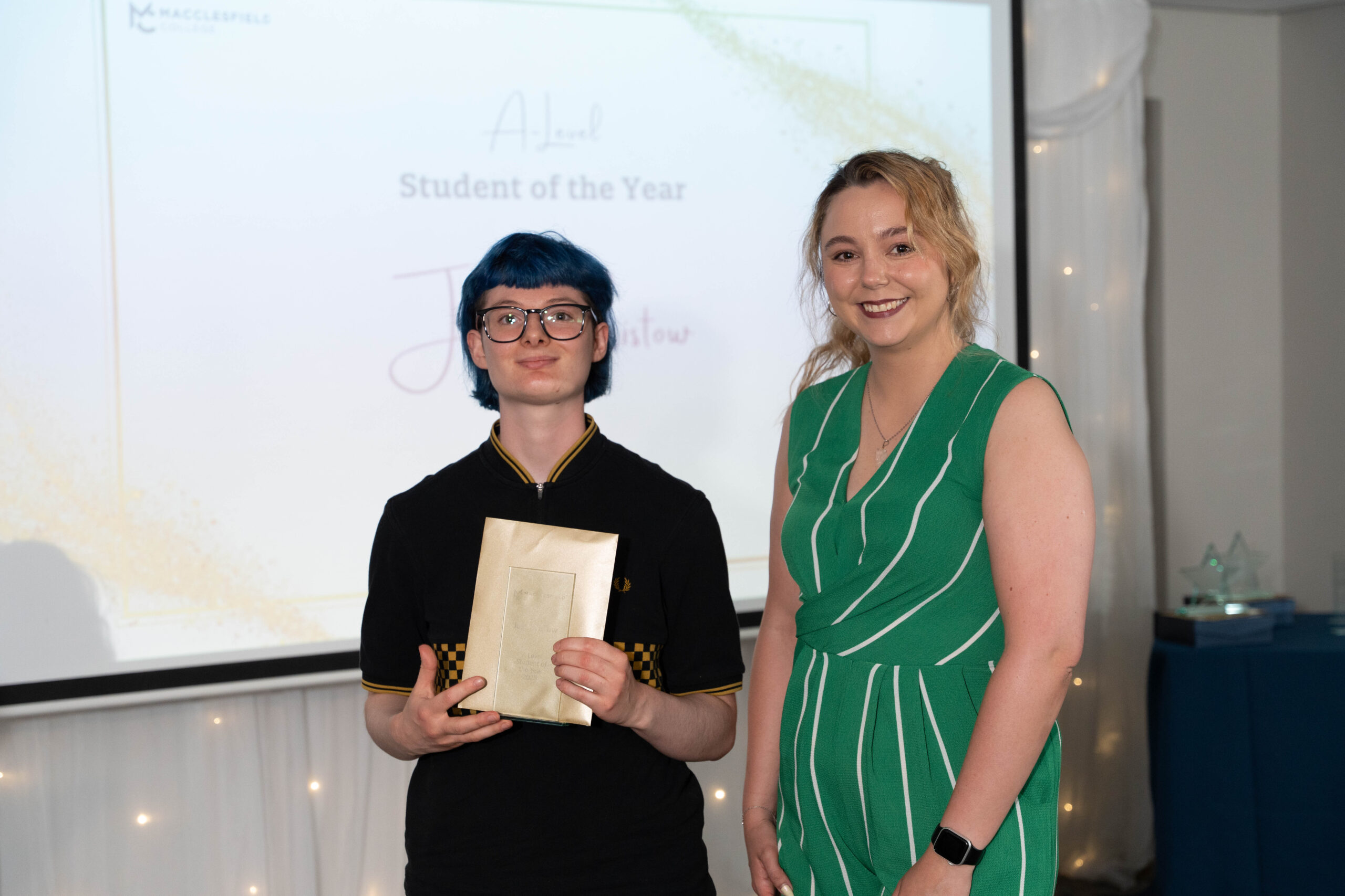 A student and their teacher are standing together, smiling for a photo, during an awards ceremony