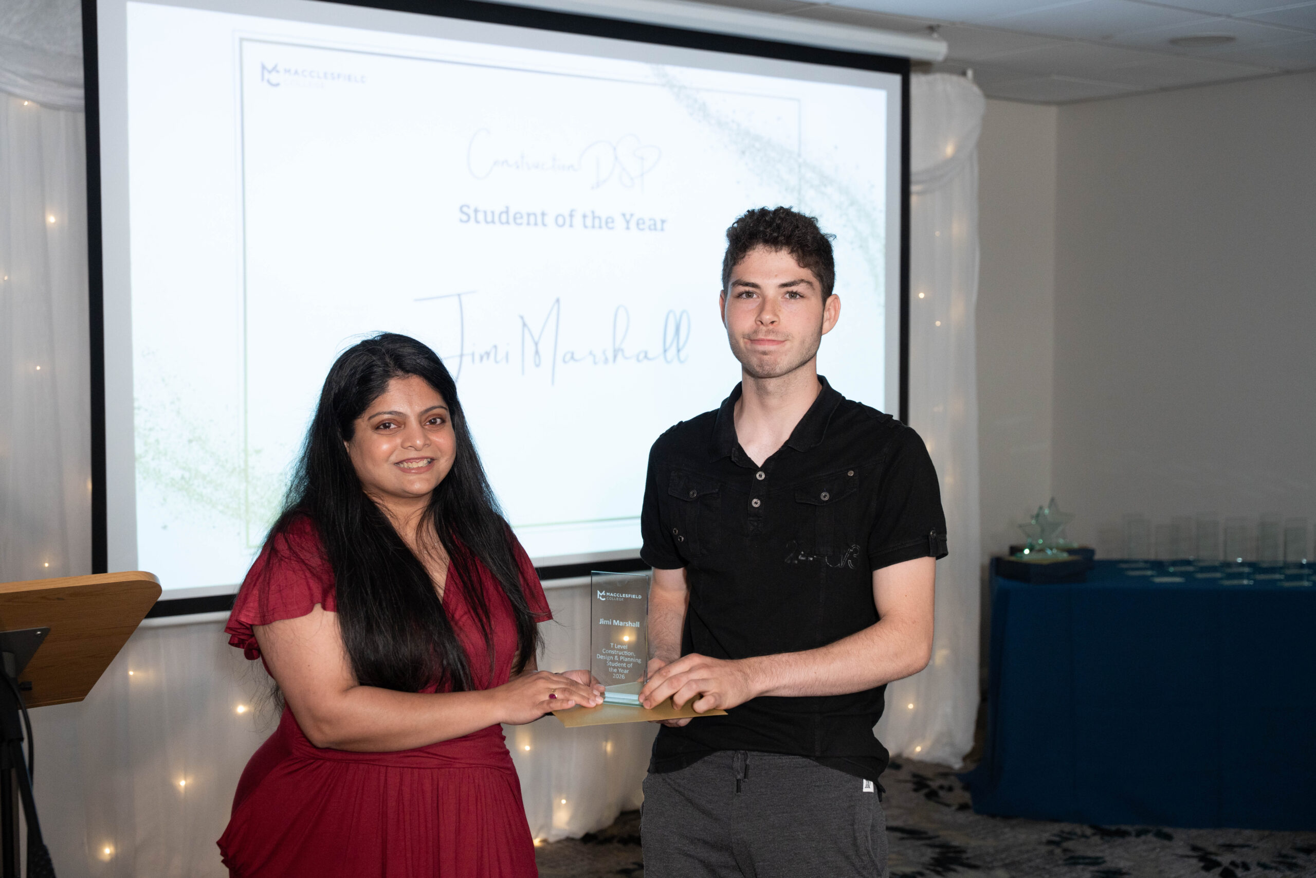 a female staff member and male student are holding a glass award and smiling for a photo, during a student awards event