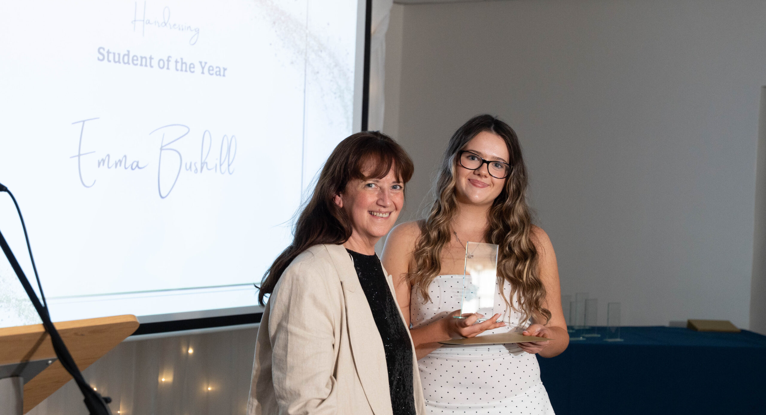 A female staff member and female student are smiling for a photo during a student awards event. The student is holding a glass award